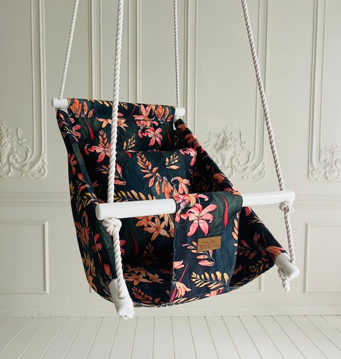 djupsinne-babygunga-hangande Handtillverkad babygunga Djupsinne med orange blommor mot mörka blad, hängande i bomullsrep mot vit vägg. Handmålade träpinnar och Videung-logga.